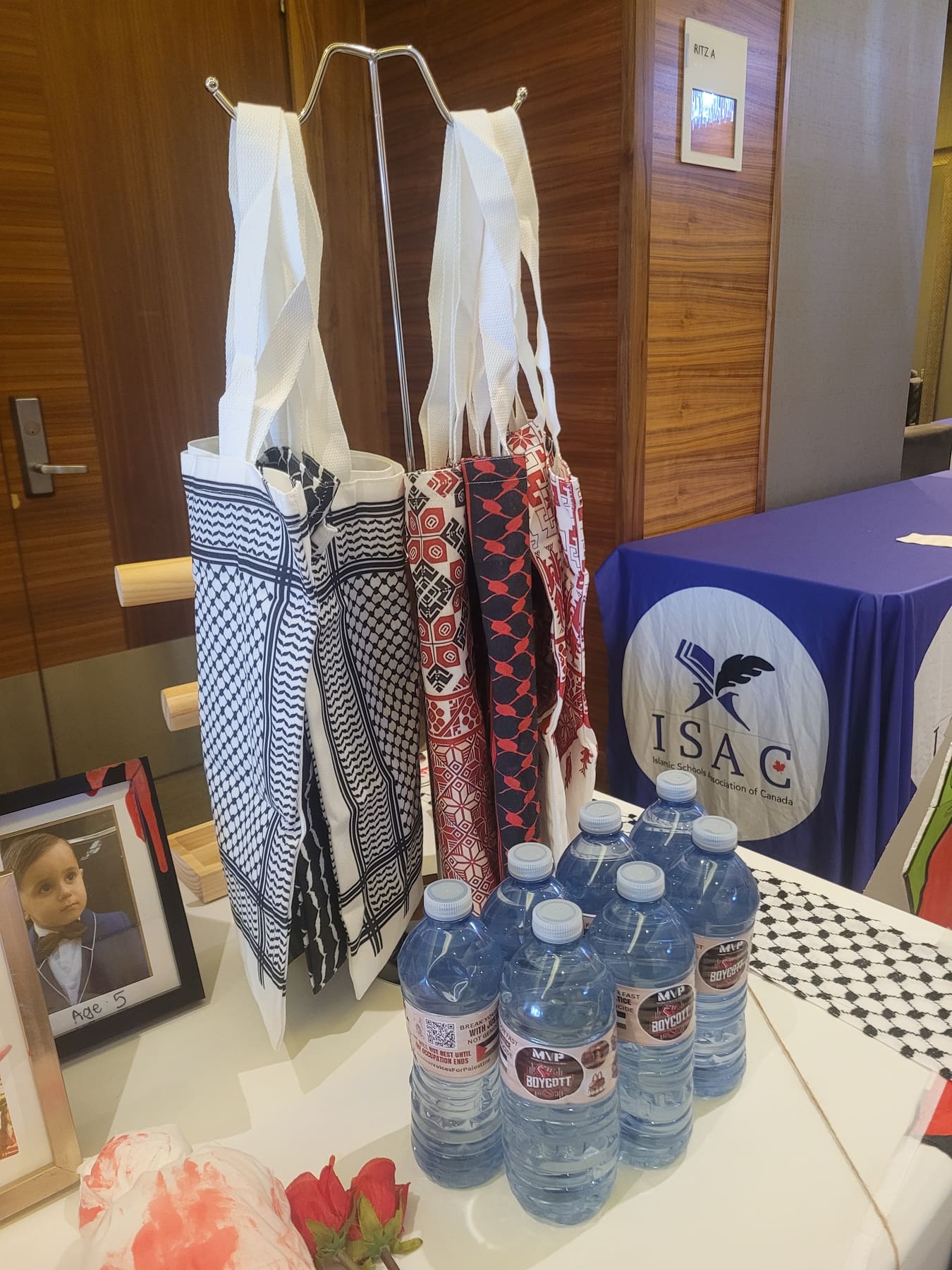 Keffiyeh-pattern tote bags hanging above MVP water bottles at a booth beside an ISAC banner