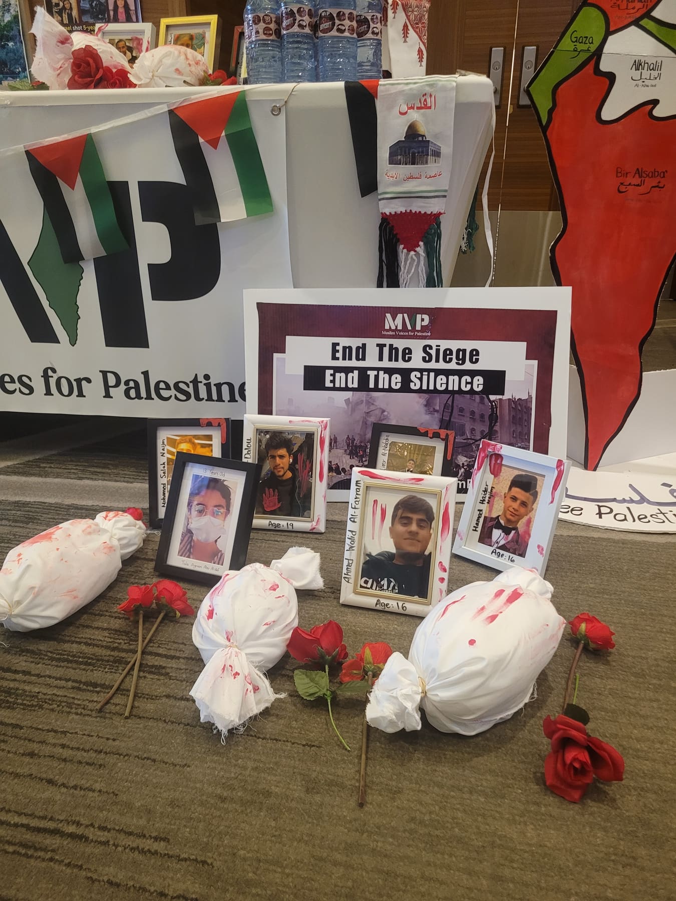A memorial of shrouded bundles, roses, and framed photographs beside an “End the Siege, End the Silence” sign