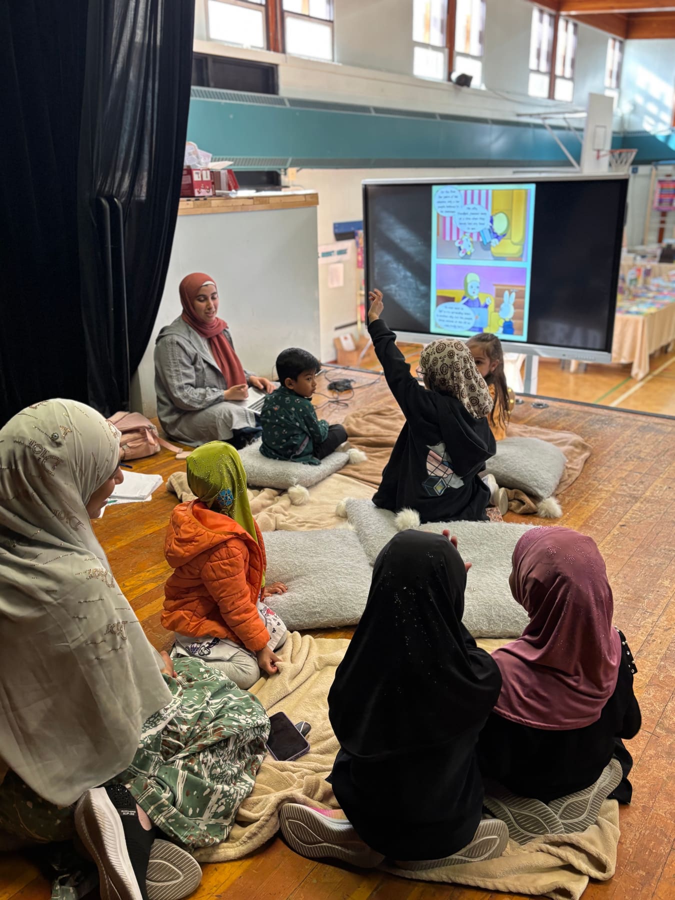 Children sitting on cushions in a gym listening to a Palestine Story Time reading shown on a screen