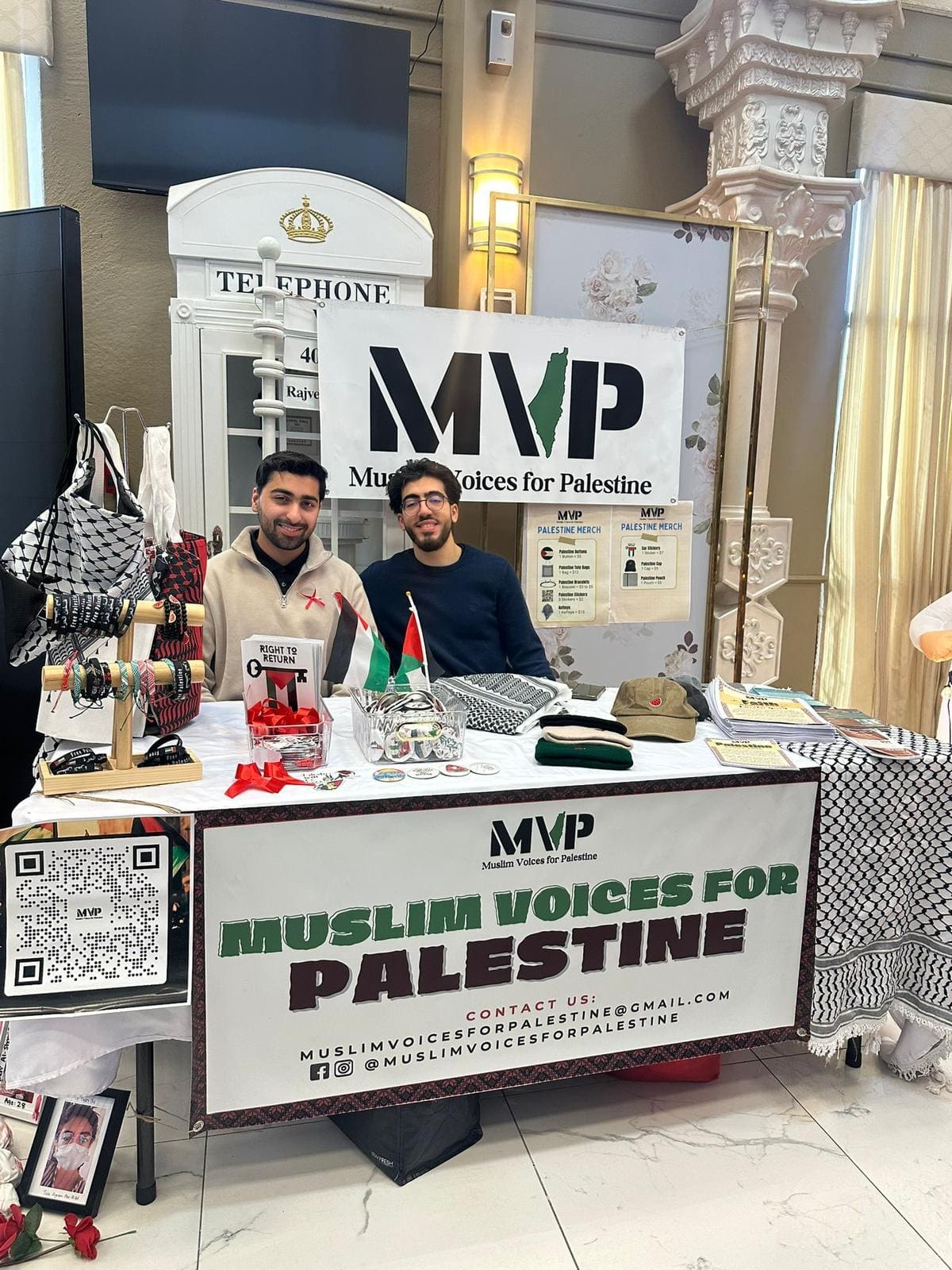 Two volunteers smiling behind the Muslim Voices for Palestine booth with keffiyehs, bracelets, and pins
